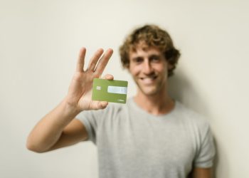 A cheerful man holding a green credit card against a neutral background.