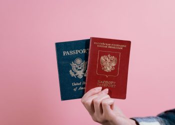 Close-up of a hand holding American and Russian passports on a pink background.